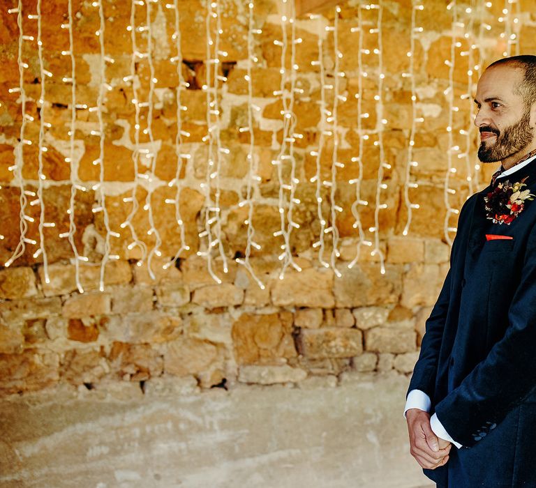 Groom in blue velvet jacket stands in front of fairy lights curtain 