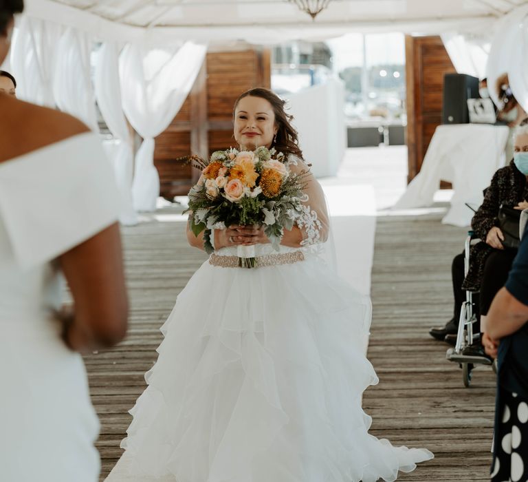 Bride in tulle skirt wedding dress walks down the aisle by herself holding an orange, peach and coral wedding bouquet 