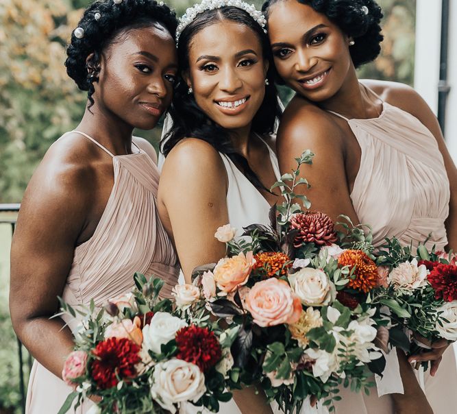 A bride stands with her bridesmaids smiling at camera. The bridesmaids wear their hair up with braids and the bride has hers half up half down. Wedding hairstyles for Black brides,