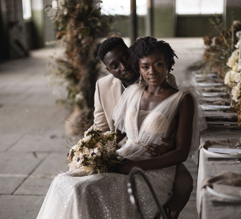 A bride and groom sit at a table. The bride is Black and wears her natural hair in small twists. She wears an embellished gown and holds and anemone bouquet.