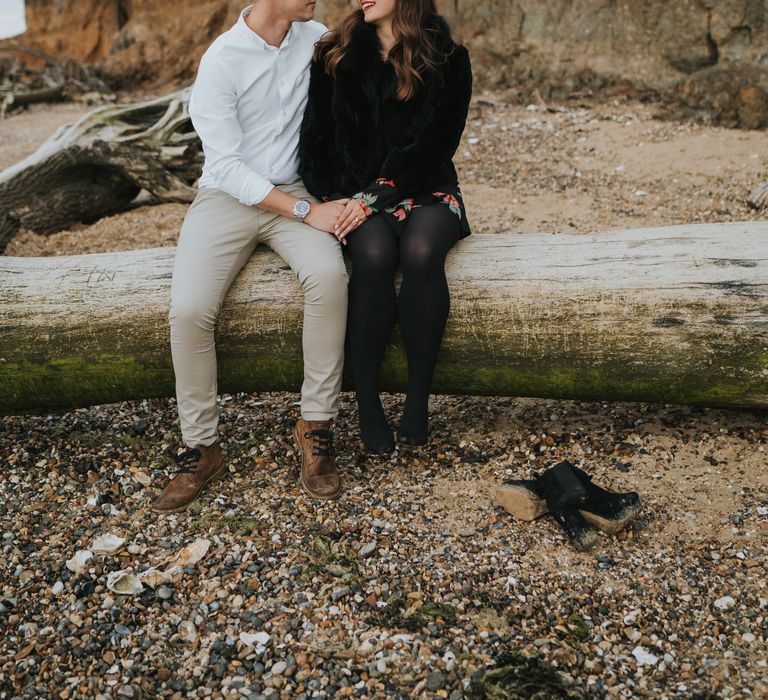 Groom-to-be in a white shirt, chino's and dessert boots sitting on a log on the beach with his Fiancé in a black and red floral dress, faux fur coat, tights and boots. 