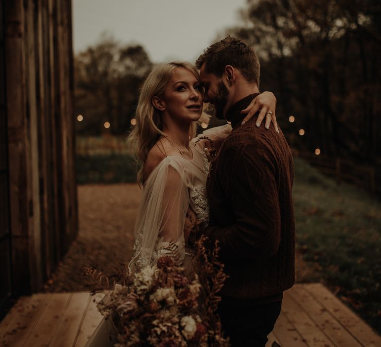 Bride with long wavy hair in a beige wedding dress embracing her groom in a brown turtle neck jumper 