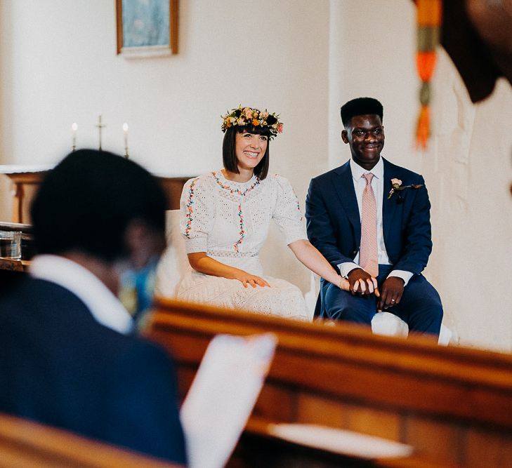 Bride in a Broderie Anglaise wedding dress resting her hand on her grooms knee during the church wedding ceremony 