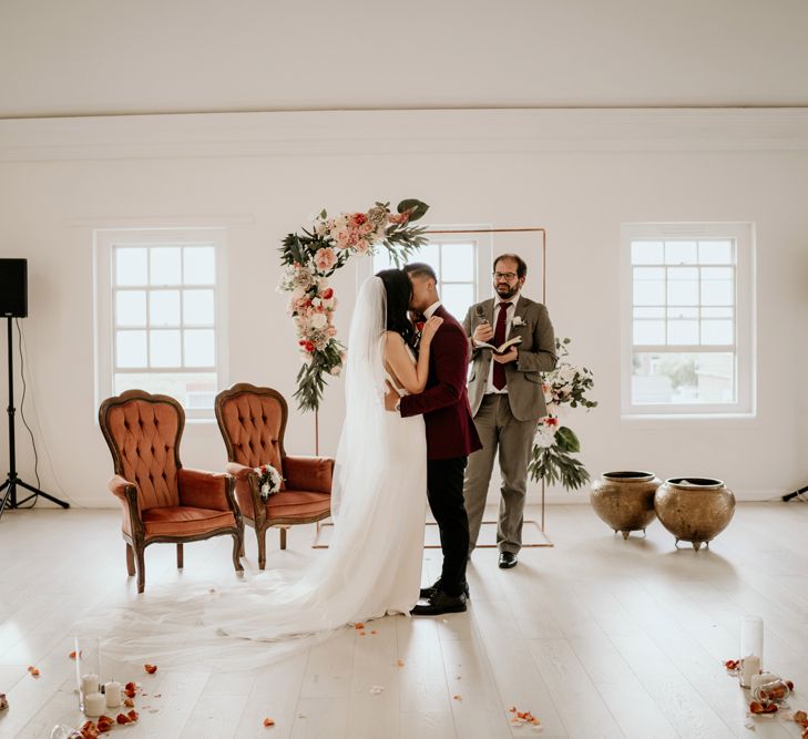 Bride and groom kissing at the altar with two velvet chairs and copper frame altar decor 