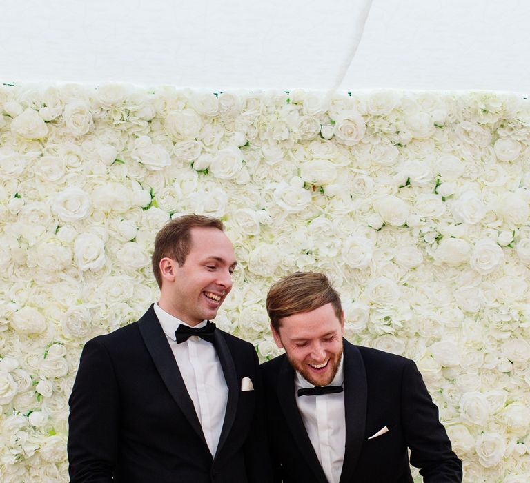 Grooms stand in front of white flower wall