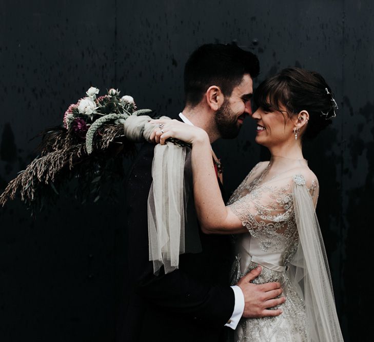 A bride and groom stand nose to nose. The brides dress has a cape and she holds a homemade wedding bouquet over the grooms shoulder. 