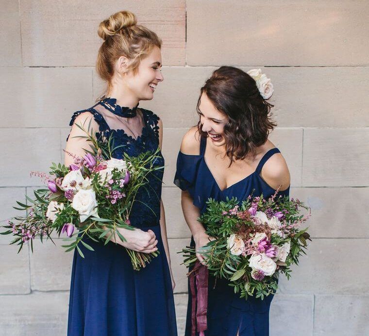 Two bridesmaids wearing similar chiffon navy bridesmaid dresses, one with a split skirt and off shoulder sleeves, the other with a sleeveless high neck mesh and lace top | Coast Dresses | Photography by WE ARE // THE CLARKES