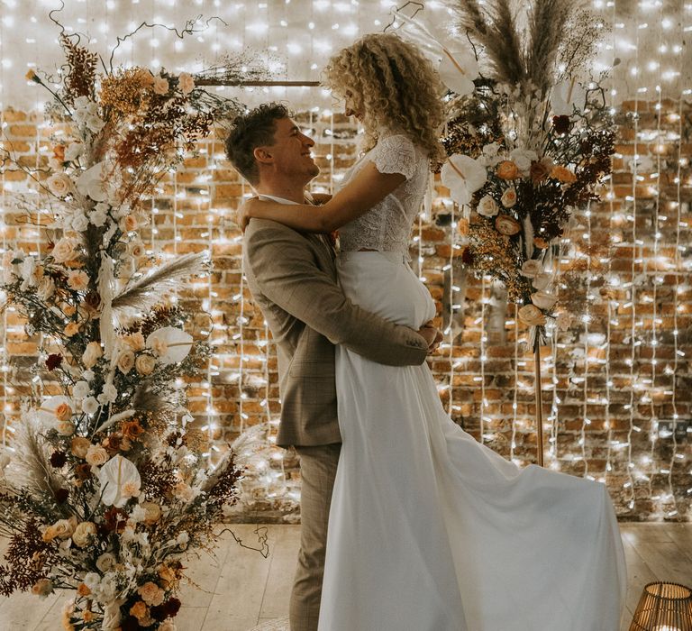 Groom lifting up blonde bride with curly wedding hair in front of altar display of neutral coloured flowers and fairy lights