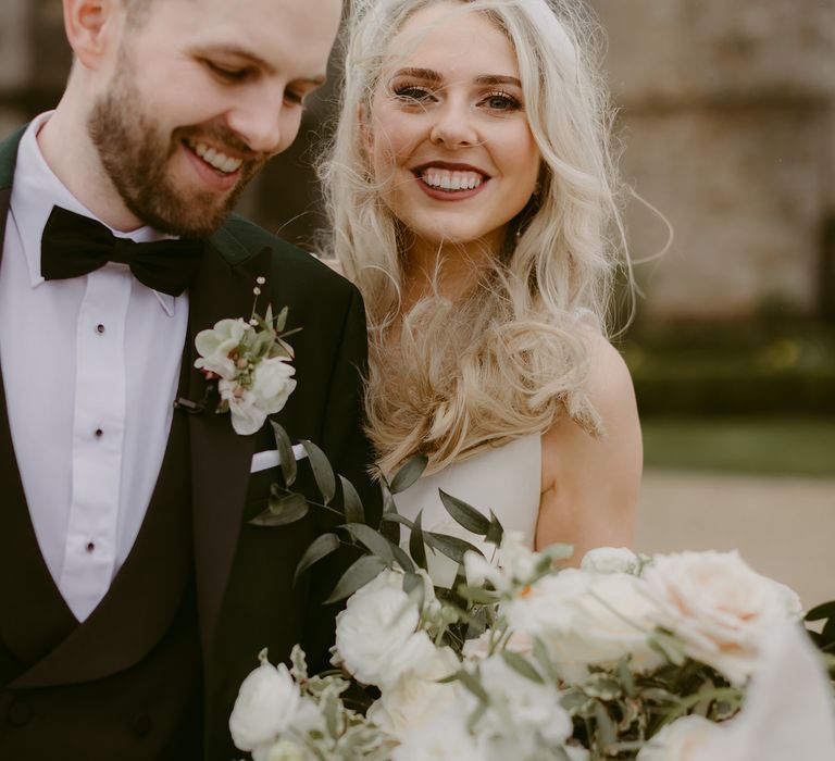 Bride & groom smile and laugh together whilst holding white floral bouquet