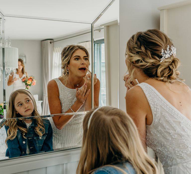 Bride applies lipstick on her wedding day in the mirror as her daughter looks on whilst wearing denim jacket