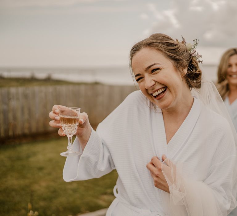 Bride in white dressing gown and veil stands in garden laughing whilst holding glass of sparkling wine at Dunluce Castle wedding