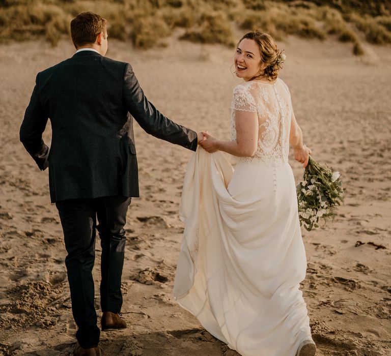 Groom in navy suit runs with bride in lace top capped sleeve wedding dress with satin skirt on the beach after Dunluce Castle wedding