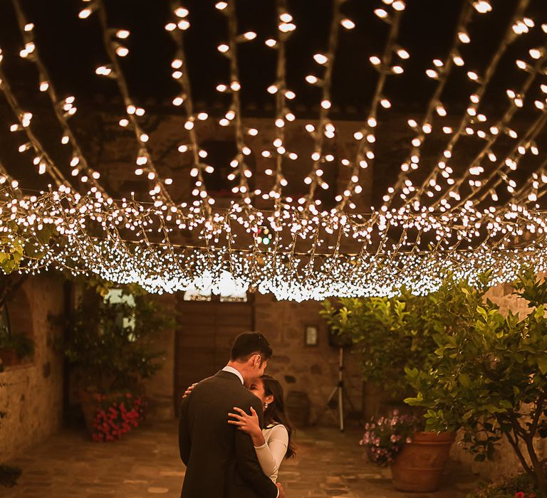 Couple stand outdoors as fairy lights shine above them during the night