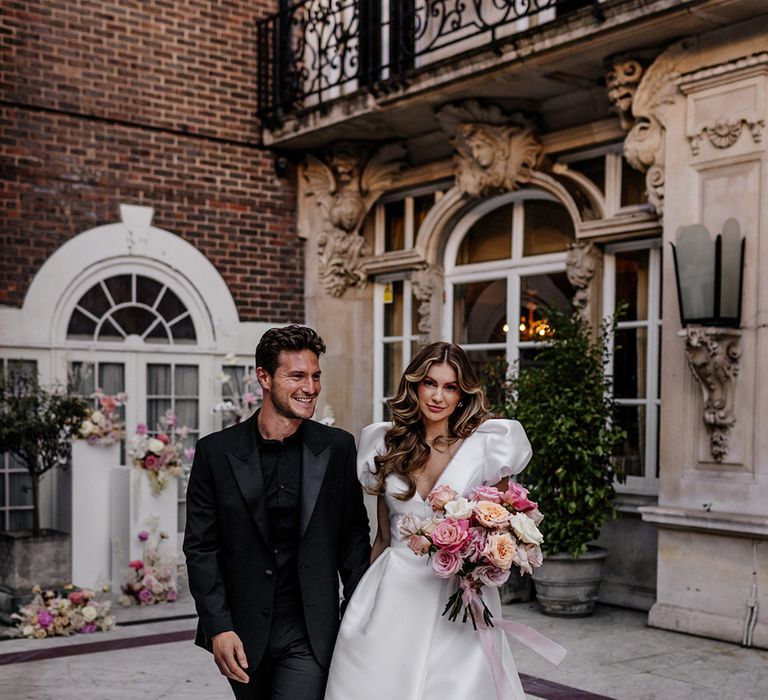 Bride in a Love in Lace bridal gown holding a pink peeled back rose wedding bouquet tied with ribbon and her husbands hand dressed in black 