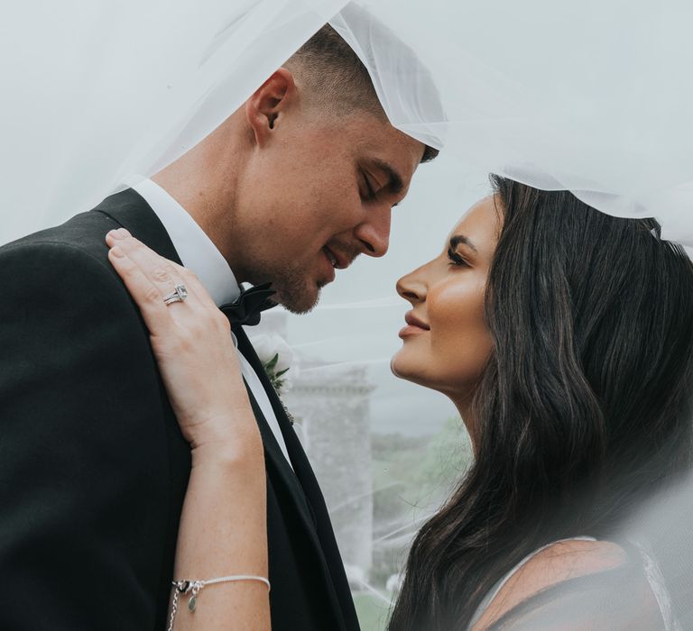 Bride with long dark curled wedding hair and groom in black tuxedo look into each others eyes underneath brides veil after their wedding at Came House Dorset 