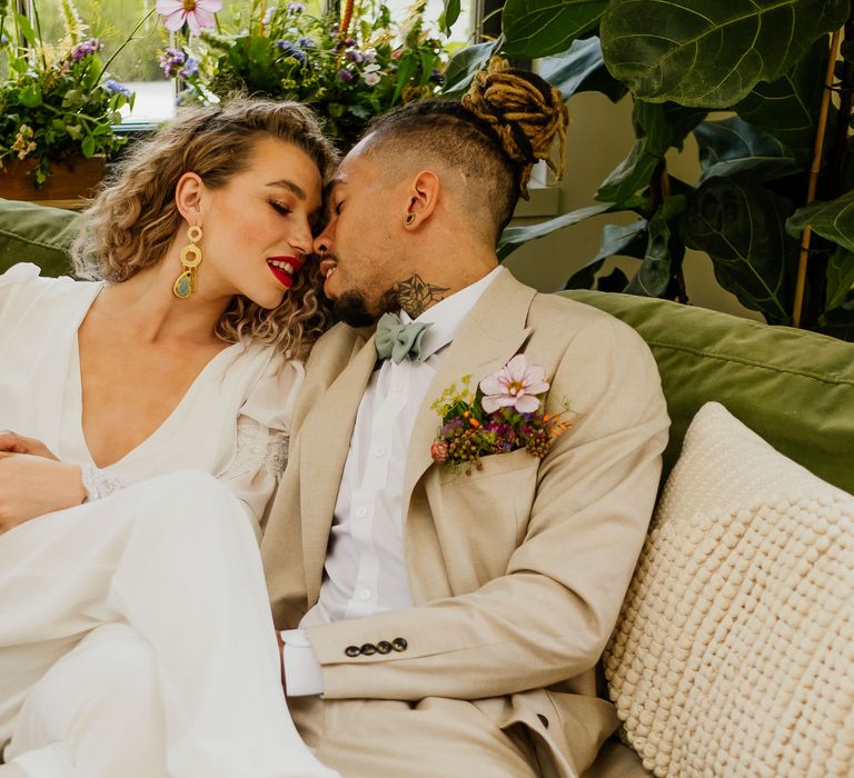 Groom in a beige suit with mint green bow tie and wildflower buttonhole flowers kissing his curly haired bride on a velvet green sofa in a bridal jumpsuit 