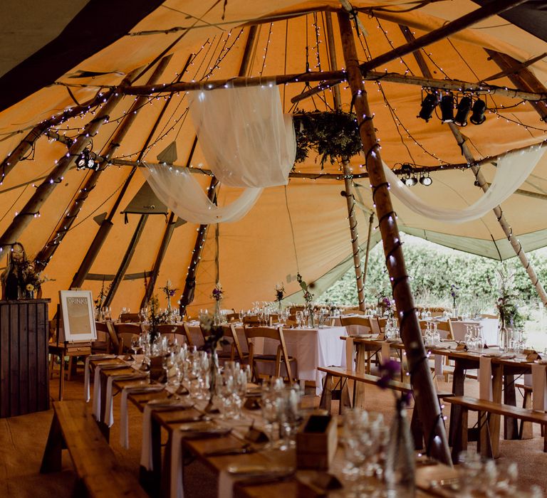 Interior of tipi wedding reception with fairy lights, draping and rustic wooden tables for home farm wedding 