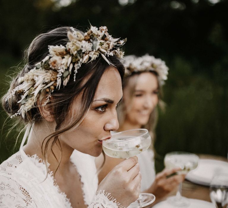 Boho bride in a lace wedding dress with long sleeves drinking a cocktail from a champagne saucer 