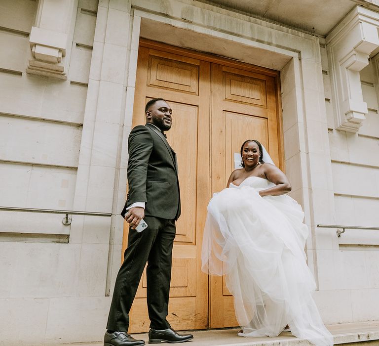 Bride & groom laugh and smile with one another as bride lifts her gown up to walk up stairs with her groom