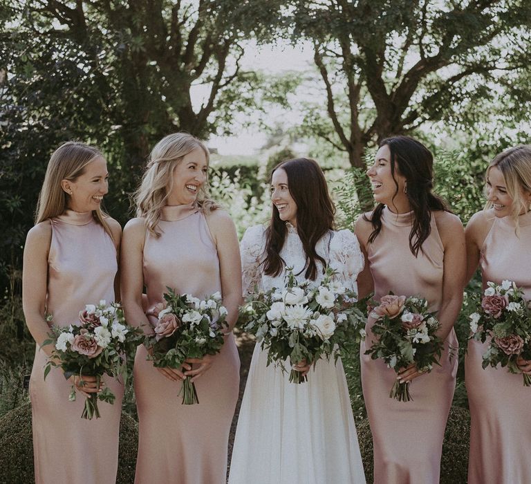 Bride in white lace puffed sleeve Daalarna wedding dress holding white and green bridal bouquet stands in the middle of four bridesmaids in blush pink satin high necked bridesmaids dresses in the grounds of Loseley Park for summer wedding