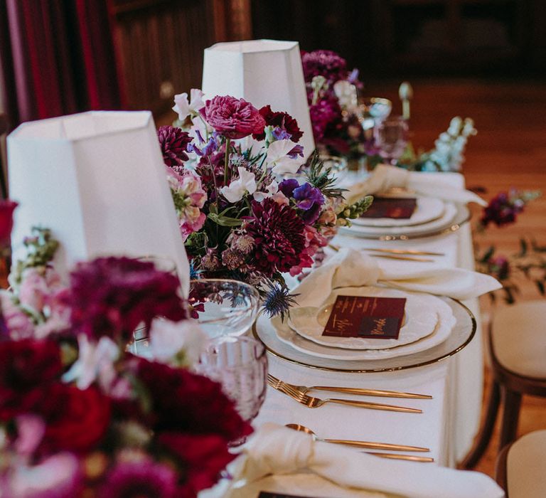 Burgundy, red, purple and pink wedding table decor with floral centrepieces, coloured glassware and gold cutlery 