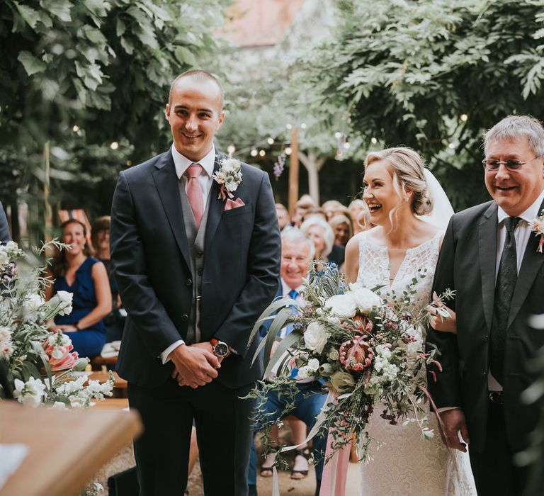 Groom in dark three piece suit with grey waistcoat and pink tie stands with hands clasped next to bride in lace Justin Alexander wedding dress holding mixed white and pink bridal bouquet arm in arm with father of the bride in black suit during outdoor wedding ceremony at Tythe Barn