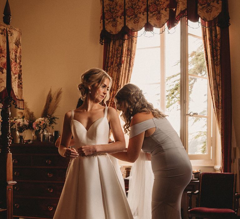 Bridesmaid in a grey fitted dress helping the bride in a Stella York A-line gown do up her wedding dress