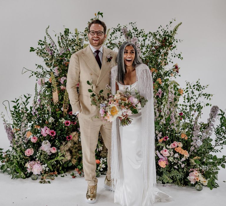 Colourful Loft Studios wedding with bride in a Halfpenny London Cape and groom in a beige suit standing at the altar in front of a colourful floral arrangement 