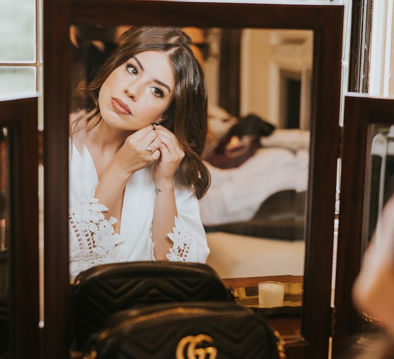 Bride in white lace trimmed bridal robe puts in her earrings as she looks in mirror at Notley Abbey wedding