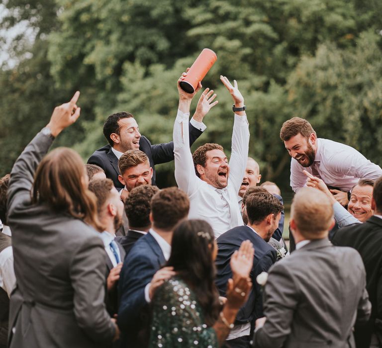 Group of wedding guests all cheer as man in white shirt and white tie catches whisky bottle box at Notley Abbey wedding