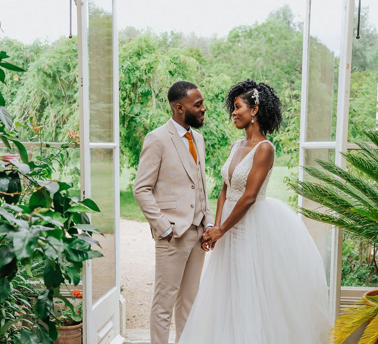 Black bride in a princess, plunging neckline wedding dress holding hands with her groom in beige suit in a conservatory at Modern Hall, London