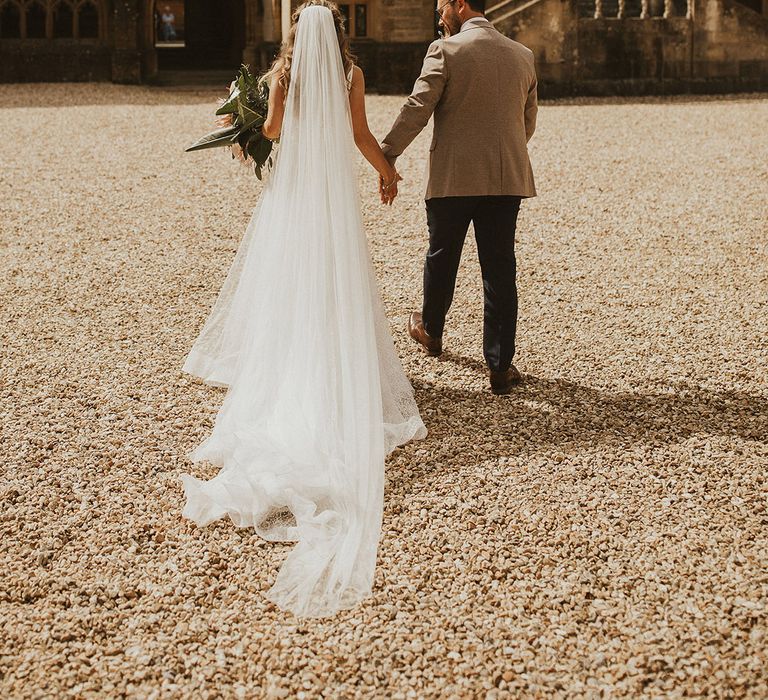 Bride & groom walk with one another outside church on their wedding day as brides veil trails behind her