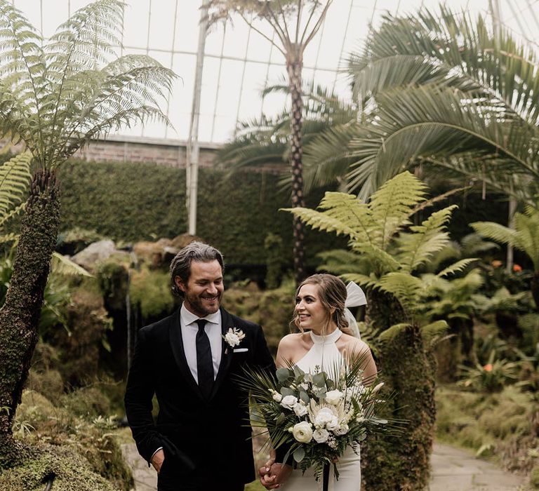 Groom in a black-tie suit and bride in a halter neck wedding dress with bow in her hair holding a white flower and green foliage wedding bouquet 