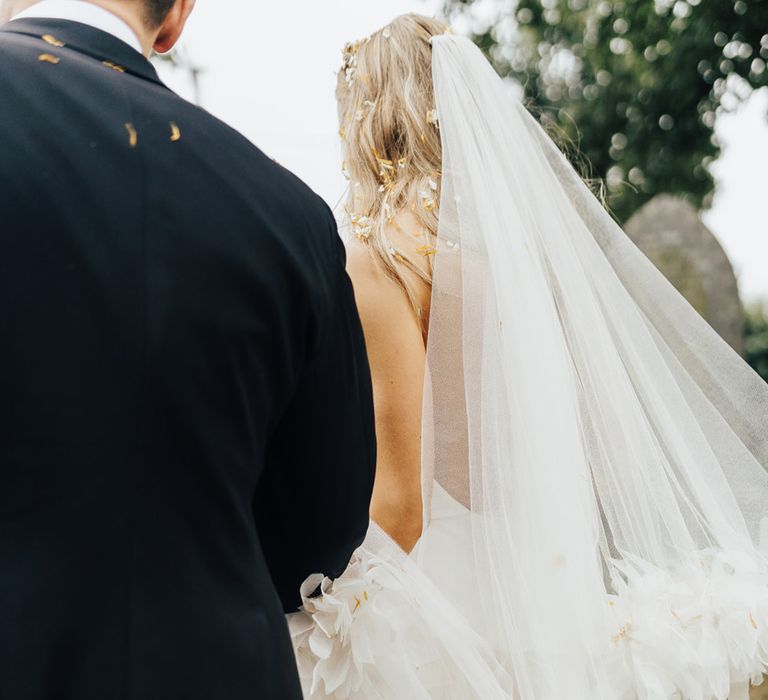 Bride in cathedral length ruffled Halfpenny London veil walks with groom in black suit across churchyard after wedding ceremony