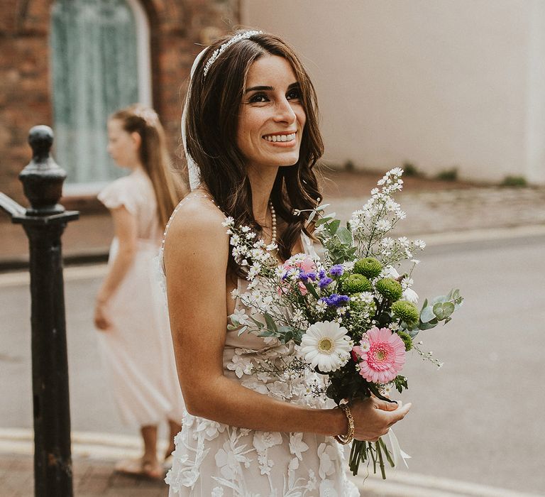 Bride smiles and holds floral bouquet and wears her brown hair down in natural curled style