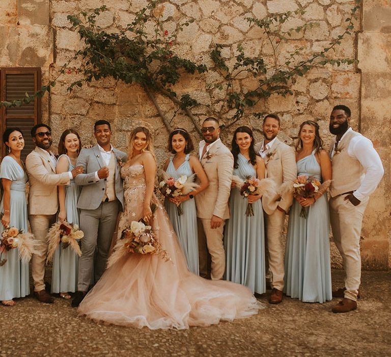 Bride & groom stand with their wedding party who wear dusty blue bridesmaid gowns and pale suits