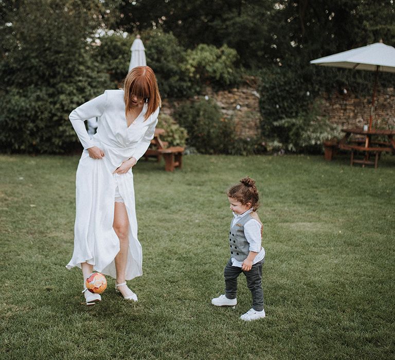 Bride in long sleeve dress plays football with little boy wedding guest