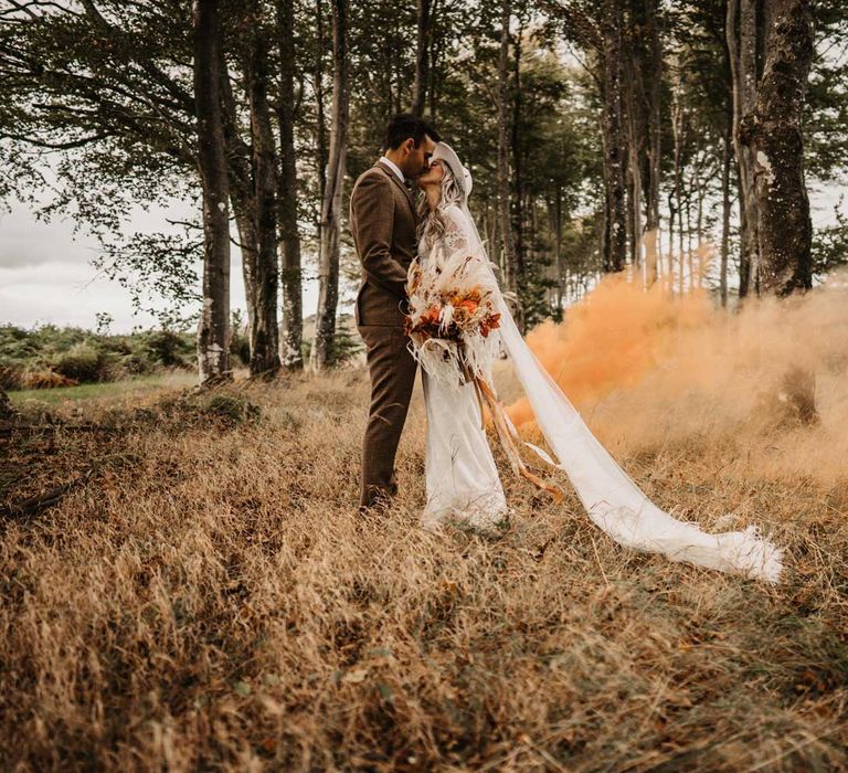 Bride in lace boho two piece wedding dress, bridal hat and veil holding white and orange boho bouquet kisses groom in brown tweed suit in woodland with orange smoke bomb in the background