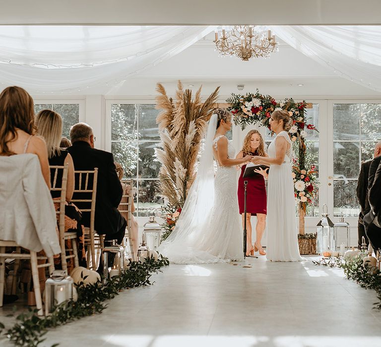 Brides hold hands in front of floral archway on their wedding day during ceremony