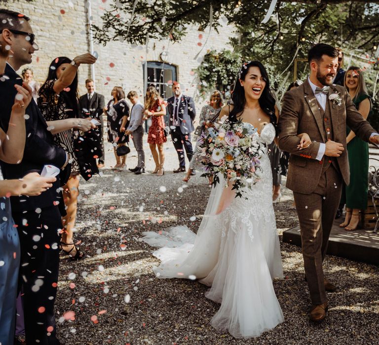 Bride in lace and tulle wedding dress holding multicoloured pastel bridal bouquet walks with groom in brown tweed suit as guests throw multicoloured confetti