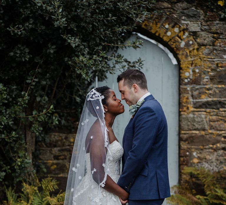 Bride wears lace edge wedding veil and embroidered detailed chiffon wedding dress as she holds hands with Groom in fitted blue suit in outdoor setting