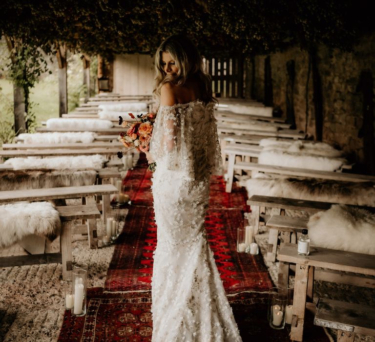 Bride in off the shoulder wedding dress with applique florals and train stands on vintage rug aisle holding orange bridal bouquet 