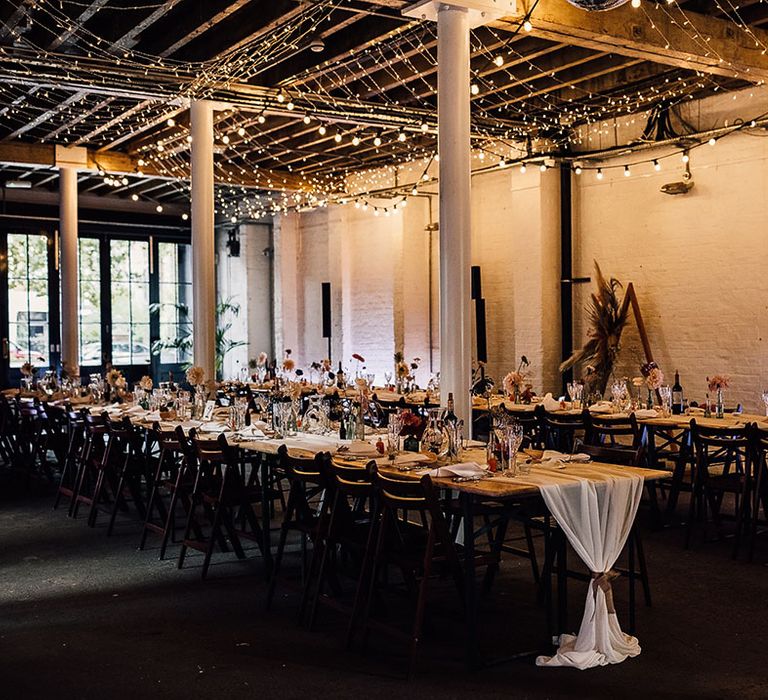 Industrial wedding reception decor at 100 Barrington with fairy light canopy, table runners and flower stems in bottles table decor 