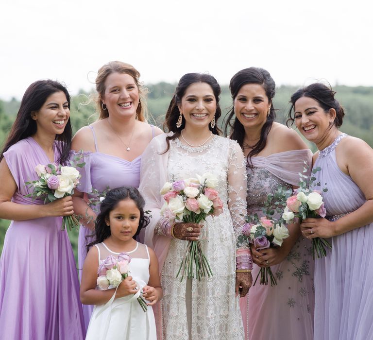 Bride stands with her bridesmaids and flower girl who wear varying colours of pastel purple whilst holding floral bouquets