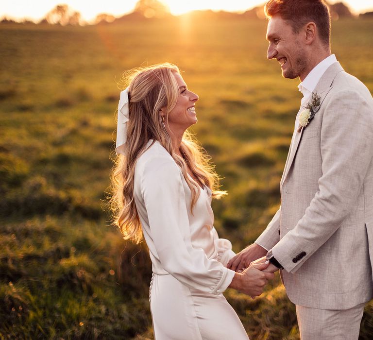 Laughing bride in long sleeve wrap top and satin bridal hair bow stands in field holding hands with groom in grey shot and yellow floral buttonhole during golden hour