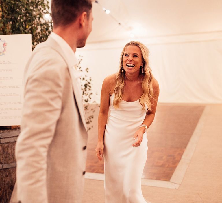 Laughing bride in satin thin strap wedding dress walks into marquee with groom in grey suit 