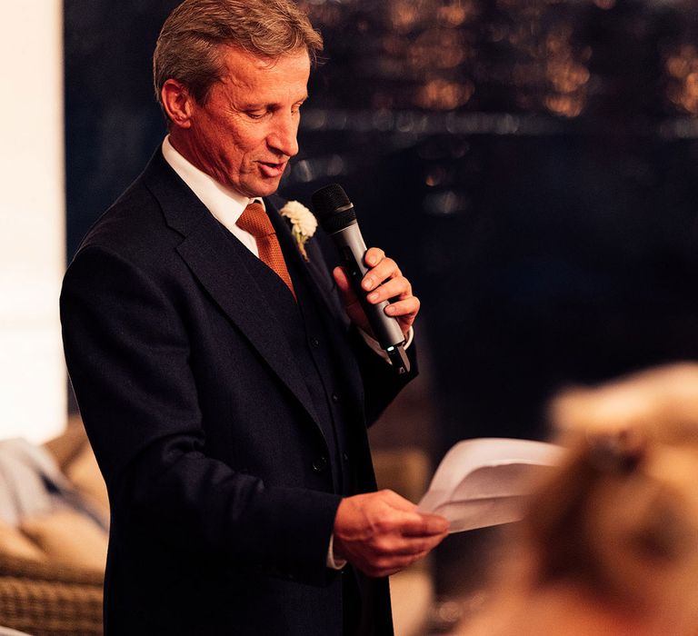 Man in blue three piece suit, orange tie and floral buttonhole holding microphone makes speech at marquee wedding reception