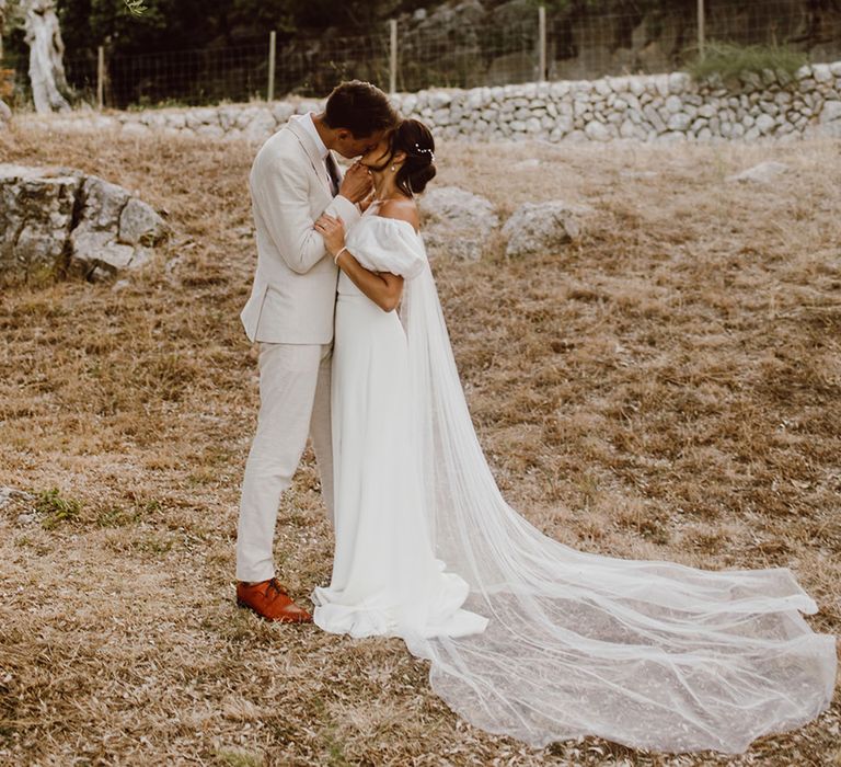 Bride in off the shoulder puff sleeve Alexandra Grecco dress shares a kiss with groom in beige suit in Mallorca