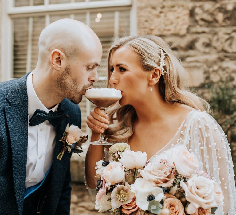 Bride with pearl detail wedding dress and pearl hair clip sits on espresso martini with groom in three piece tweed suit with bow tie