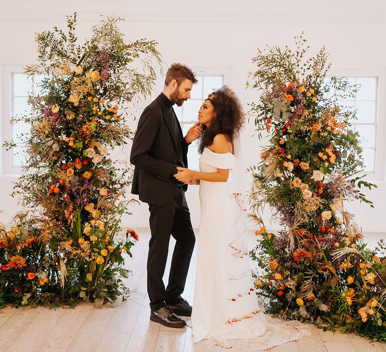 Groom in all-black touching his brides face in an off the shoulder wedding dress in-between orange and green column wedding ceremony flower arrangements 